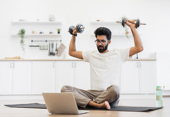 Young man sitting on mat exercising with dumbbells, recording health video blog indoors. Scene...