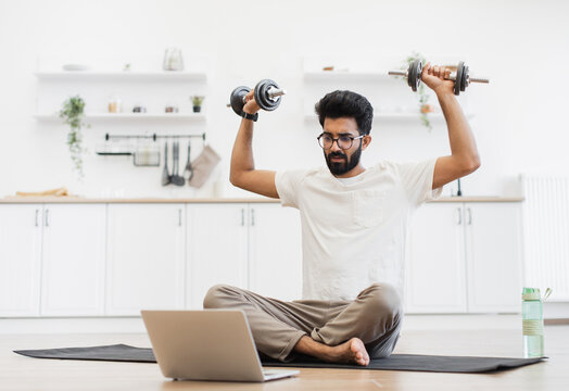 Young man sitting on mat exercising with dumbbells, recording health video blog indoors. Scene suggests fitness, technology, and home routine concept. - Powered by Adobe