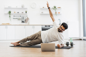 Young adult man in his 30s performs fitness side plank in bright minimalist kitchen using yoga mat, laptop, dumbbells, and water bottle for workout and video blogging in home fitness routine.