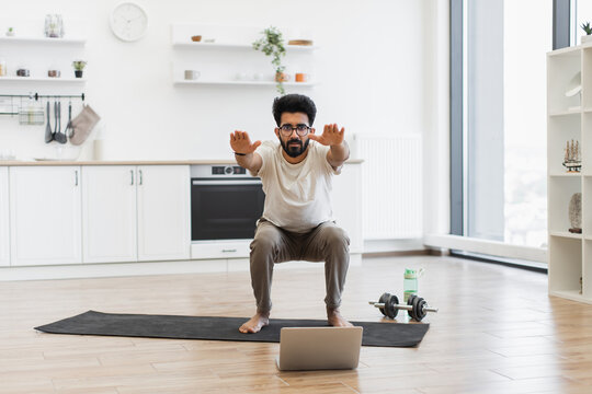 Young man performing squats in bright home kitchen using laptop for virtual fitness routine. Demonstrates home fitness, active lifestyle. Background shows well-lit modern household environment