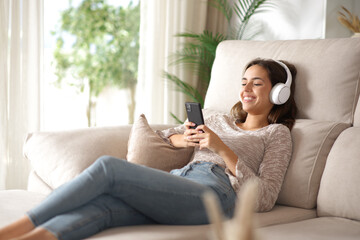 Happy woman using phone and headphones on a sofa