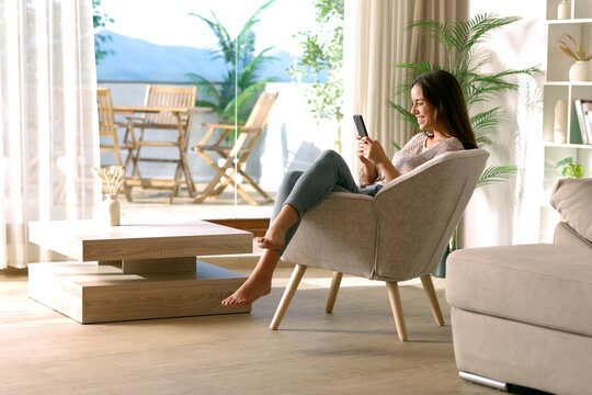 Happy woman using phone sitting on armchair at home