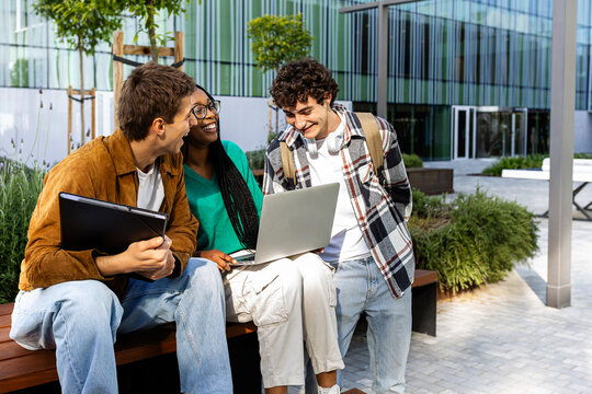 University students working on project outdoors with laptop and tablet, sharing ideas and smiling