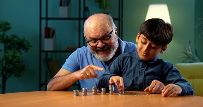 Indian grandfather and kid making coins tower at home, elderly man teaching grandson value of money, inculcating savings habit for better financial future, family bonding moment indoors