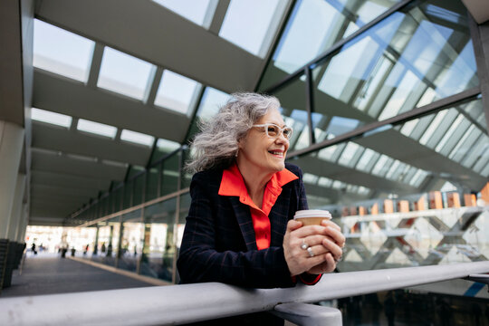 Happy mature businesswoman with disposable coffee cup leaning on railing