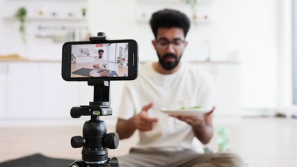 Young adult male with beard records video blog in home kitchen holding salad bowl, discussing healthy eating habits and sharing tips. Scene shows content creation, cooking, modern lifestyle concepts.