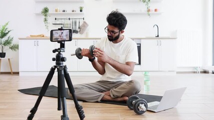 Man sitting on yoga mat recording fitness blog at home using dumbbell and tripod camera setup. Concept highlights health, fitness, and modern lifestyle promoting home-recorded fitness video