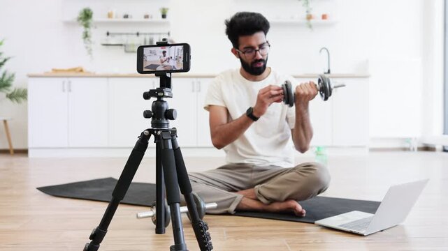 Adult man records fitness video blog at household kitchen, explaining dumbbell exercises. Sitting on floor with camera, tripod, and laptop, demonstrating healthy lifestyle ideas.