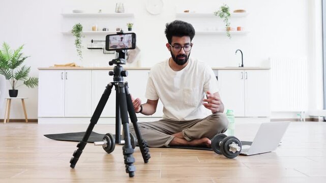 Man exercising with dumbbells during video recording in kitchen, showcasing fitness routine and health. Seated on mat near laptop and phone, promoting home fitness motivation.