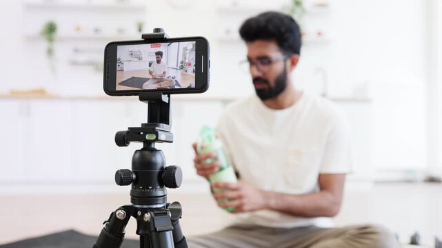 Man in his 30s filming fitness vlog in home kitchen holding water bottle, concept of staying hydrated during workouts, sports, and daily fitness routines, emphasizing health and well-being