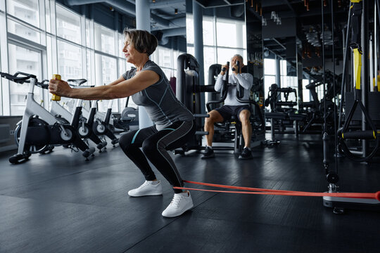Mature woman doing squats with a rubber band with exercising man on the background