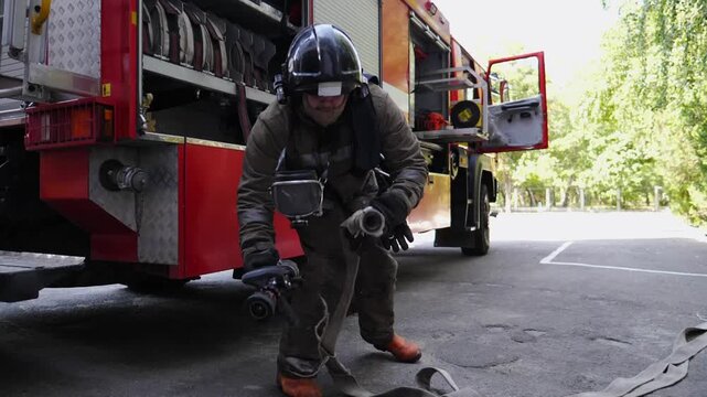 Professional fireman in full equipment connecting fire hydrant to hose near big red truck. Young male fireguard getting ready to extinguish fire. Saving lives and heroic profession concept. Slow mo