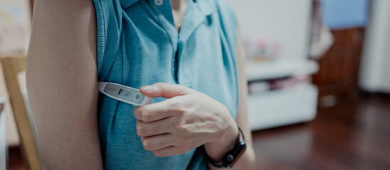 A person checking body temperature with a digital thermometer under the armpit, screen showing measurement, concept of health monitoring, medical diagnosis, illness prevention, and daily care.