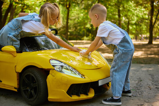 Children washing toy yellow car with sponge and foam in summer park. Concept of responsibility, playful childhood activity, outdoor teamwork, and positive learning. - Powered by Adobe