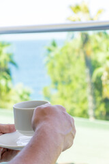 Close-up of hand holding coffee cup on balcony with panoramic view of tropical greenery, palm trees, and sea horizon. Perfect summer vacation scene representing relaxation, lifestyle, mindfulness, and