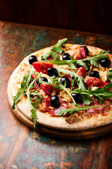 Pizza with salami, tomatoes and mozzarella cheese, black olives and arugula. Rustic wooden background. Soft focus.