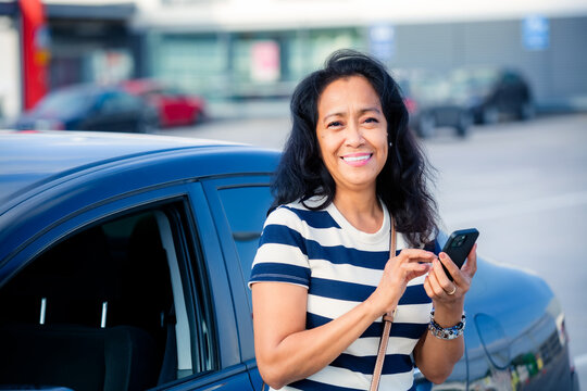 Middle-aged Asian woman stands by her car, using a smartphone, looking at camera, showing modern business, connection, and everyday urban life. - Powered by Adobe