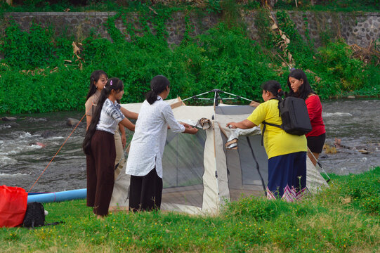 Five teenage girls cooperate to roll up the curtain and neaten their riverside tent, ensuring a proper camping setup while laughing and working together on green grass.