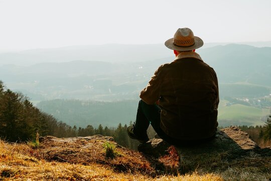 Middle Eastern man in a hat sitting on a rock enjoying a scenic mountain view during a sunny day - Powered by Adobe
