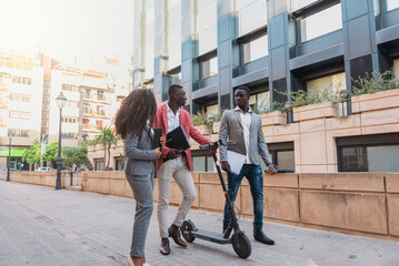 Three multiracial business people walking and talking on a city street with electric scooter