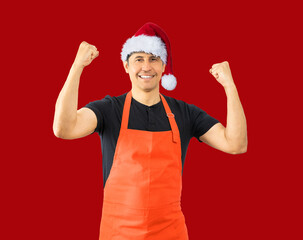 Store employee and red apron standing over isolated red background happy doing winner gesture with arms raised, smiling and screaming for success. Celebration concept while wearing a Christmas hat