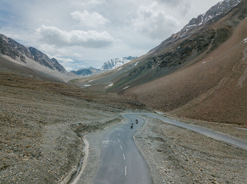 Aerial view of a winding asphalt road snaking through the rugged, arid mountains, contrasting with patches of snow under a cloudy sky, Kaza, Himachal Pradesh, India.