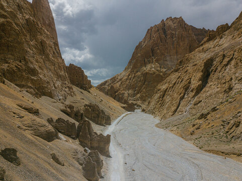 Aerial view of stark, sun-baked cliffs embracing a winding riverbed under a dramatic sky in the rugged terrain of Zanskar, Ladakh, India.