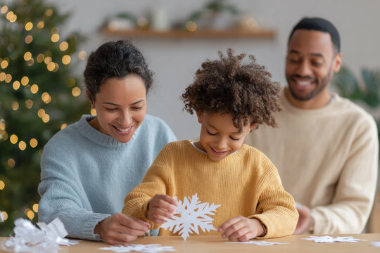 Happy African American family making Christmas craft together at home. Loving parents watch their child create beautiful paper snowflake for holiday season celebration - Powered by Adobe