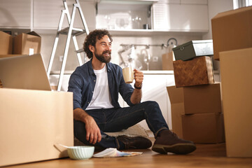 A man sits on the floor surrounded by moving boxes, smiling and holding a cup while taking a break during the busy packing process in his new kitchen.