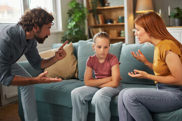 Parents express frustration in a heated argument, while their child sits quietly on the sofa, appearing upset by the ongoing conflict at home.