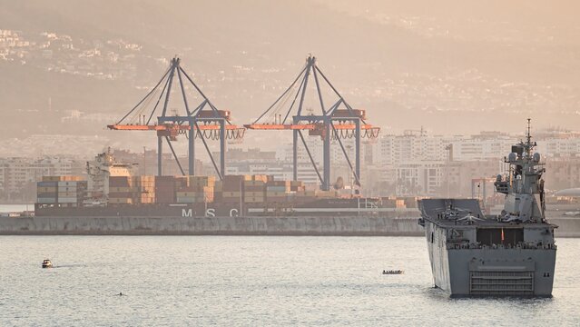 Military ship and container vessel in busy seaport at sunset, with giant cranes and city skyline in the background