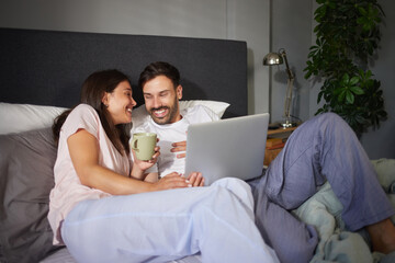 A couple shares a light moment as they plan their vacation in bed, sipping coffee and using a laptop together. The warm and inviting atmosphere enhances their connection and excitement. © Stockphotodirectors