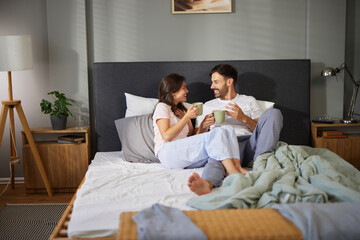A couple sits on a neatly made bed, smiling and sharing a conversation while sipping coffee. The soft morning light creates a warm ambiance in their cozy bedroom.