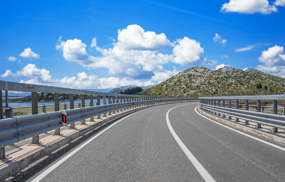 Scenic coastal highway with a curve and guardrails overlooking mountains, blue sky, and water on a bright summer day.