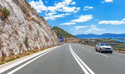A silver SUV drives along a winding highway next to a rocky hillside with a scenic coastal and mountain landscape under a bright blue sky with scattered clouds.