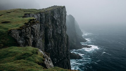 Misty cliff face overlooking turbulent ocean. Lush green vegetation clings to the rugged edge of a dramatic, grey cliff, meeting a churning, dark-blue ocean