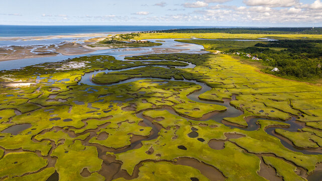Aerial view of winding waterways carve through vibrant green marshes, leading to the sandy shores and the vast ocean beyond, Cape Cod, Massachusetts, United States.