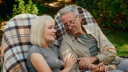 In a peaceful garden, an older couple shares a tender conversation while sitting comfortably on oversized chairs. Sunlight casts a warm glow on their joyful expressions and surroundings.