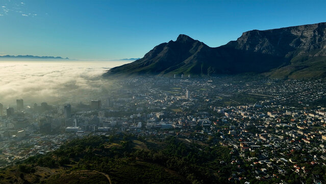 Aerial view of Table Mountain looming over Cape Town, with a blanket of clouds obscuring parts of the city, creating a dramatic contrast of light and shadow, Cape Town, Western Cape, South Africa.