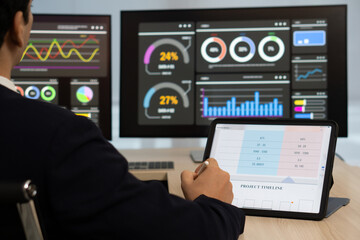 Fototapeta premium A business analyst sit at a desk, focus on data analysis using a tablet, while several monitor display various digital dashboard with colorful graph and statistic.