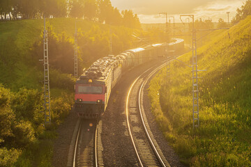 Freight train and railway. A locomotive with freight wagons and oil tanks rides among the hills at sunset