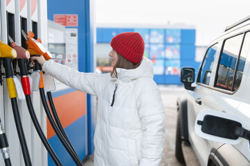 A woman in a white jacket takes off a fuel dispenser gun at a gas station. She was going to fill up her car with gasoline. Gasoline and diesel prices at gas stations. © Vladimir Razgulyaev