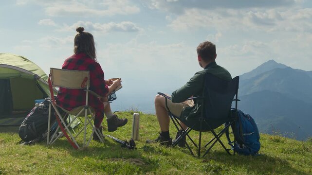 Camper couple sitting at their mountain campsite, pointing toward distant peaks, enjoying a breathtaking panoramic view of mountains and clear sky, rear view.
