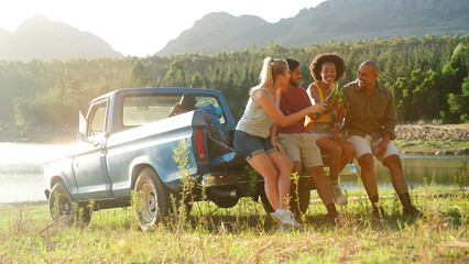 Friends With Backpacks Sitting On Tailgate Of Pick Up Truck On Road Trip By Lake Drinking Beer