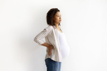 Thoughtful African American pregnant woman holding back while standing. White background portrait, symbol of prenatal discomfort and maternity challenges, copy space