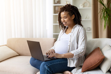 Happy pregnant African American woman sits on sofa working on laptop at home. Remote work and modern prenatal lifestyle concept casual outfit, positive mood, digital connection