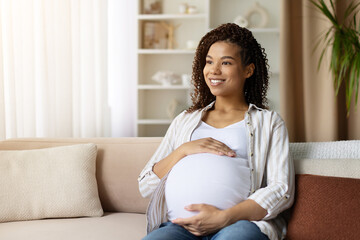 Smiling pregnant woman relaxes on sofa with hands on belly. Bright home interior, symbol of prenatal joy, love, and peaceful maternity lifestyle, copy space