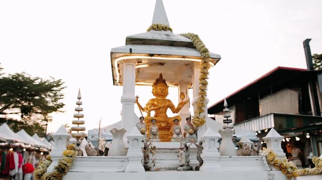 Golden brahma statue, surrounded by smaller figures and offerings, sits under an ornate white canopy in thailand.
