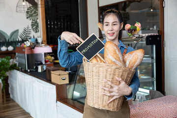 Young barista in denim apron holding a large basket of fresh baguettes with a promotion sign inside a modern bakery café, showcasing marketing strategy, product display, and small business