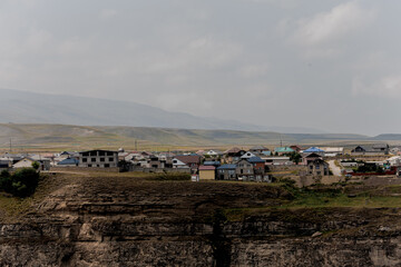 A panoramic view of a Caucasian village nestled in a mountainous landscape. The scene features traditional houses and a rugged terrain under a cloudy sky.